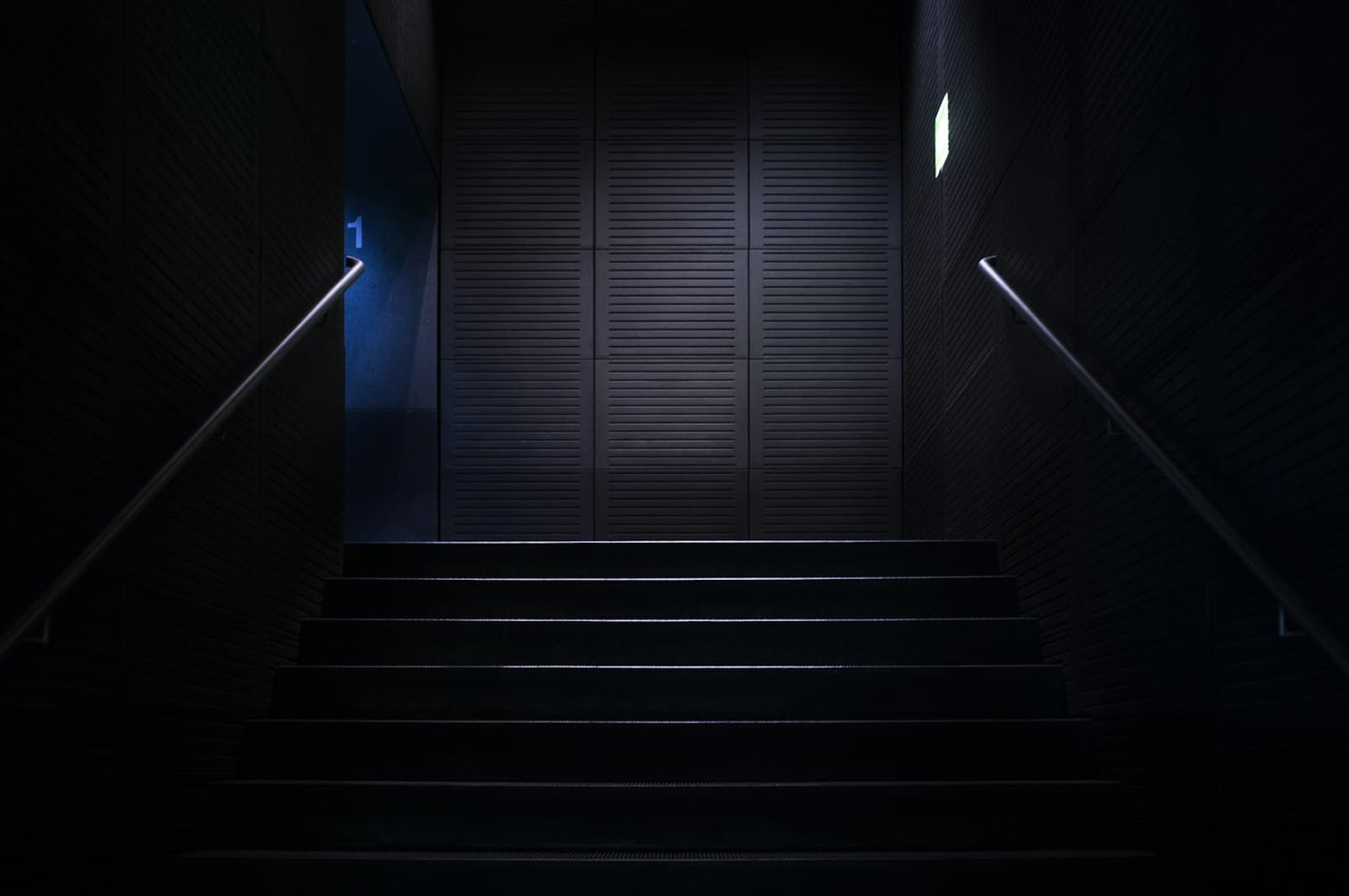 Volée de marches sombres montant vers un palier éclairé, rampes métalliques de part et d'autre.